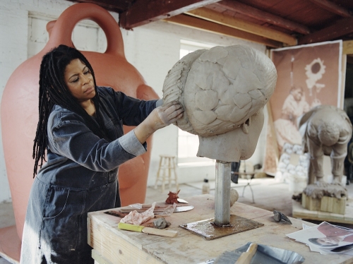 Simone Leigh, a medium-dark-skinned woman with dreadlocks in a denim work outfit, works on a large clay sculpture. 