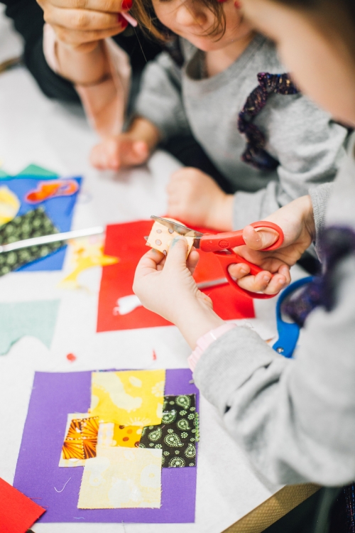 Two youths working on their art project at a table while one is cutting a piece of fabric.