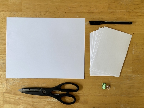 Activity supplies on a wood table which includes index cards, letter size paper, pen, scissors, and a binder clip.