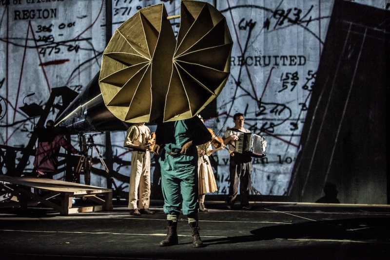 A person stands onstage with a large bell-shaped form, possibly of cardboard, covering their face. Behind them are several musicians and a backdrop showing drawings and diagrams.