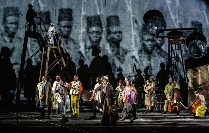 About a dozen African musicians onstage in colorful clothing. The backdrop is an archival black-and-white photograph of Africans in uniform with fez-like hats. 