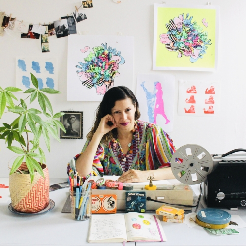 A woman with long brown hair smiles at her desk with artmaking materials displayed and small drawings hanging in the background