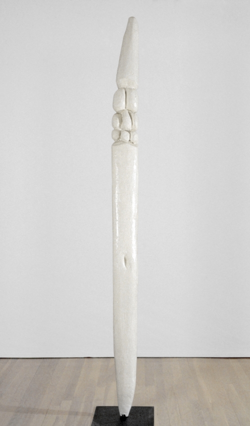 A tall, slender sculpture in white wood of an abstracted figure with carved rounded shapes at the top and a navel-like depression near the midpoint.