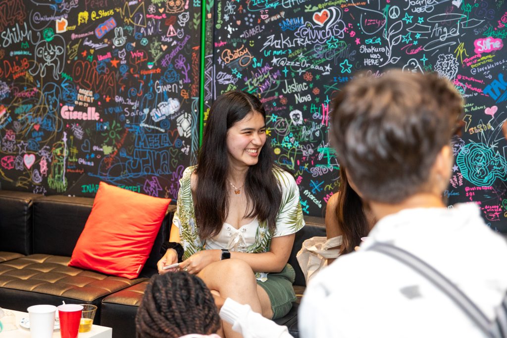 A young woman smiles in front of a black wall covered with written names and doodles.