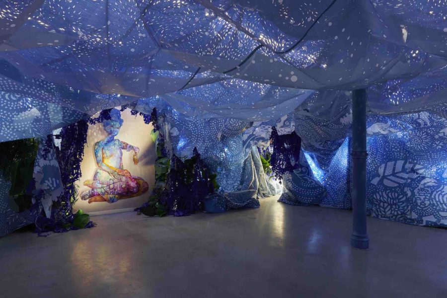 A room with artistic fabric draped from the ceiling, creating a blue, ethereal atmosphere. A colorful illuminated painting of a seated figure is displayed on the left wall. The space is filled with shadows and light patterns.